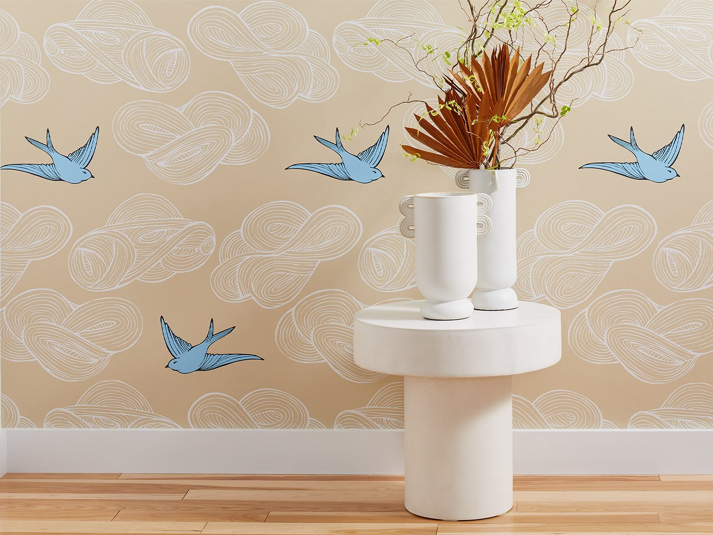 A wallpapered wall featuring a tan background with white clouds and blue birds. A white table sits in front with two white vases, one containing a dry floral arrangement