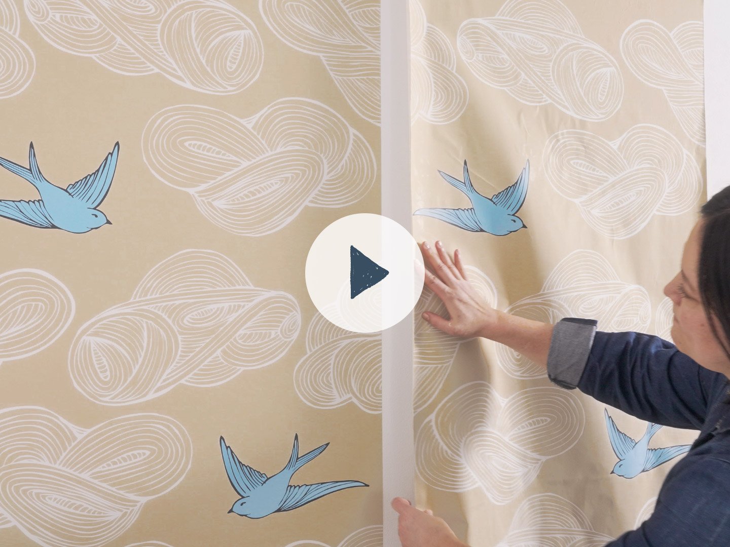 A woman with black hair positioning a strip of tan wallpaper with white clouds and blue birds on a wall next to another strip of the same wallpaper already adhering to the wall. An illustrated play button is overlaid on the image.