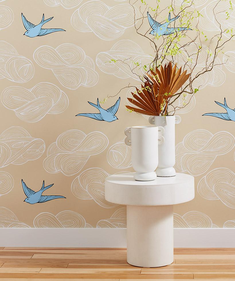 A wallpapered wall featuring a tan background with white clouds and blue birds. A white table sits in front with two white vases, one containing a dry floral arrangement